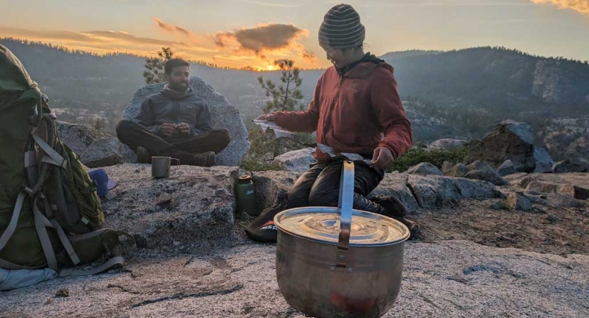 A person kneels beside a cooking pot, while another sits on a rock nearby. Behind them, the sun sets over a mountain ridge. 